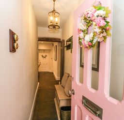 A hallway with a pink front door and floral wreath and a wooden bench with cushions at Pear Tree Cottage in Grange Over Sands