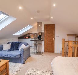 A small living area with a blue sofa, kitchenette with bar stools, wooden railing and bed at The Halt in Maiden Newton