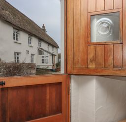 A half-open wooden door with a small glass window showing a thatched roof cottage outside at Cherryblossom Cottage in Winkleigh