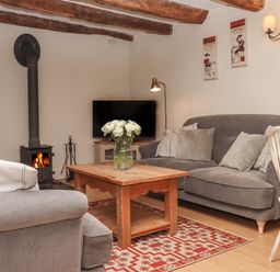 A living room with a wood stove a wooden coffee table with flowers a gray sofa and armchair a television and wooden ceiling beams at Cherryblossom Cottage in Winkleigh