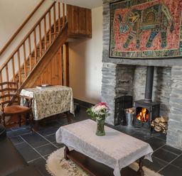 A living room with a stone fireplace and wood burning stove a wooden staircase a table with a floral tablecloth and a chair at Ty Llugwy in Capel Curig near Betws-Y-Coed