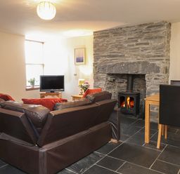 A living room with a brown leather sofa red cushions stone fireplace wood stove television and wooden dining table with black chairs at Ty Llewelyn in Capel Curig near Betws Y Coed