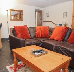 A living room with a brown leather sofa and orange cushions a wooden coffee table a wooden dining table with black chairs a floor lamp a wall mirror and wooden door at Ty Llewelyn in Capel Curig near Betws-Y-Coed