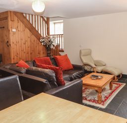 A sitting area with a black leather sofa with red cushions a wooden coffee table on a rug and a beige armchair with an ottoman near wooden stairs at Ty Llewelyn in Capel Curig near Betws-Y-Coed