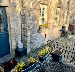 A stone building exterior with a blue door a small black table with a snowman figure flowerpots with plants a fenced stone patio with a round glass table and stacked black chairs at 1 Boarbank Farm Allithwaite near Cartmel