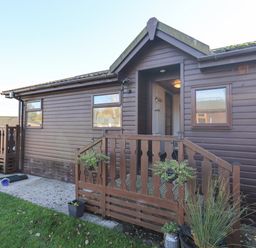 The exterior of a single-story wooden house with two windows and a small fenced porch with hanging plants at Kellet Lodge Lakeside 26 Dock Acres near Carnforth
