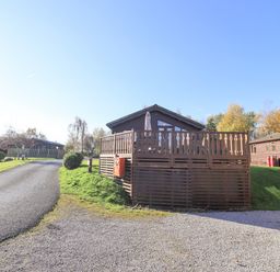 A wooden lodge with a fenced porch next to a paved road and grassy area at Kellet Lodge Lakeside 26 in Dock Acres near Carnforth