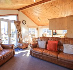 A living room with leather sofas and cushions near glass doors leading to a deck with outdoor furniture at Kellet Lodge Lakeside 26 Dock Acres near Carnforth