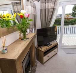 A living room with a wooden fireplace mantel with flowers and reed diffusers a flat screen tv on a wooden stand and sliding glass doors leading to a deck at 117 Crosswinds Hillway near Bembridge