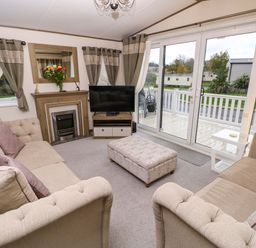 A living room with two sofas a patterned ottoman a fireplace a TV on a stand and large sliding glass doors at 117 Crosswinds Hillway near Bembridge