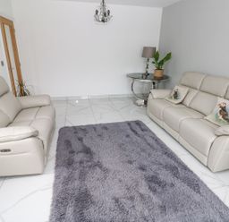 A living room with two beige sofas a round side table with a lamp and plant and a gray rug on white tiled floor at Golygfa Hafan in Little Haven