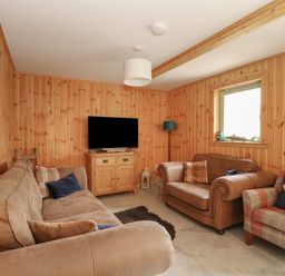A living room with wooden panel walls a television on a wooden cabinet a brown sectional sofa two armchairs and a floor lamp at Bradan Lodge in Dalmally