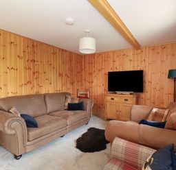 A living room with wooden panel walls two brown sofas a plaid armchair a television on a wooden cabinet a floor lamp and a small rug on the floor at Bradan Lodge in Dalmally