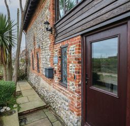 The exterior of a stone and brick house with a wooden door and windows and a pathway with plants at Bridge Barn in Sidestrand near Overstrand