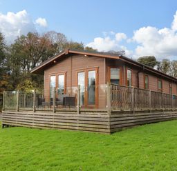 A brown wooden lodge with glass and wooden railing on a green lawn surrounded by trees at Lakeview Lodge in Forton near Garstang