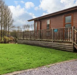 A cabin with a wooden deck and railing overlooking a grassy yard and trees at Lakeview Lodge in Forton near Garstang