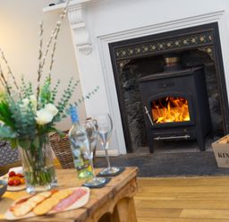 A living room with a wood burning stove with fire a wooden table with snacks wine bottle and glasses and a vase with flowers at Captains Walk in Newquay