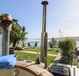 An outdoor seating area with a wooden hot tub a bottle of champagne two glasses and chairs overlooking a body of water at Fronwig New Quay