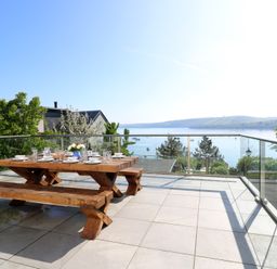 An outdoor patio with a wooden picnic table and benches set for dining overlooking a body of water with hills in the background at Fronwig in New Quay