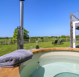 An outdoor hot tub with a towel a bottle of champagne and glasses on the edge and lawn chairs on grass at Yr Adfa in Llangeitho near Tregaron