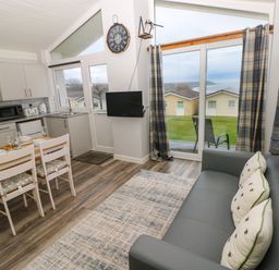 A kitchen and dining area with a table set for four next to a gray sofa and large sliding glass doors showing outdoor cabins at Chalet 22 in Port Eynon