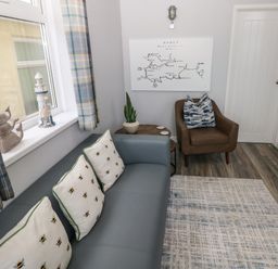 A seating area with a blue sofa with bee patterned cushions a brown armchair a small round wooden table a potted plant a wall map and a window with plaid curtains at Chalet 22 in Port Eynon