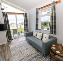 A living room with a gray sofa and patterned cushions a round wooden table with a plant a wall clock and a television at Chalet 22 in Port Eynon