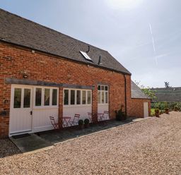 Exterior view of a brick building with white framed windows and doors with red tables and chairs outside at Home Farm Barn in Todenham near Moreton-In-Marsh