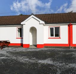 A white bungalow with red trim and a brown tiled roof with a wooden picnic table in front at Robins Roost 2 in Cornamona County Galway