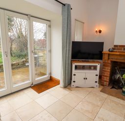 A room with tiled floor glass doors leading outside a television on a white stand and a brick fireplace at Christon Well Cottage in Christon Bank near Embleton