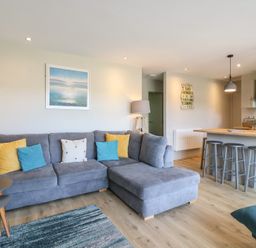 A living room with a gray sectional sofa and colorful cushions near a kitchen island with three stools at 1 Kessingland Cottages in Kessingland