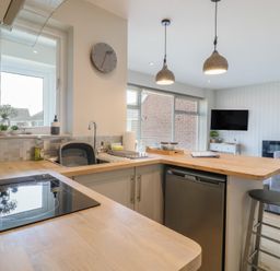 A kitchen with wooden countertops a sink a stove and hanging lights next to a living area with a sofa and a wall mounted TV at 1 Kessingland Cottages in Kessingland