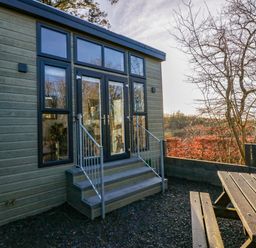 A small wooden building with glass doors and windows next to a picnic table outdoors at Elderflower Lodge in Allithwaite near Cartmel