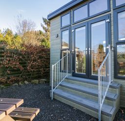 Exterior view of a building with steps leading to glass doors and a gravel ground with a wooden picnic table nearby at Elderflower Lodge in Allithwaite near Cartmel