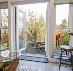A room with glass doors leading to an outdoor area with a wooden picnic table and metal stove at Elderflower Lodge in Allithwaite near Cartmel