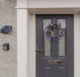 A front door with two small windows and a wreath with purple flowers next to a wall plaque reading 1 Brookside in Warcop