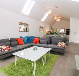 A living area with a gray sectional sofa with colorful pillows and blankets a white coffee table on a green rug and a dining area in the background at Ty Chwarel in Menai Bridge