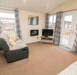 A living room with two sofas a wall mounted fireplace a TV on a stand and windows with patterned curtains at 6 Sherwood in Carnforth near Tewitfield