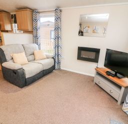 A living room with a sofa a television on a stand and a wall-mounted electric fireplace at 6 Sherwood in Carnforth near Tewitfield