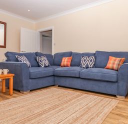 A living room with a blue sectional sofa with patterned and plaid cushions wooden side tables with mugs and a remote and a large beige rug on a wooden floor at West View in Northlew near Hatherleigh