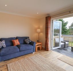 A living room with a blue sofa and cushions a floor lamp wooden side tables and an open door leading to an outdoor patio with wicker chairs at West View in Northlew near Hatherleigh