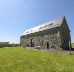 An exterior view of a stone barn with a pitched roof and skylights next to a grassy lawn and a wooden fence at Tithe Barn in Capel Mawr near Malltraeth