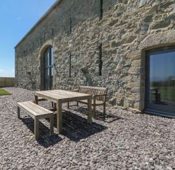 An outdoor patio with wooden table bench and chair on gravel beside a stone building at Tithe Barn in Capel Mawr near Malltraeth