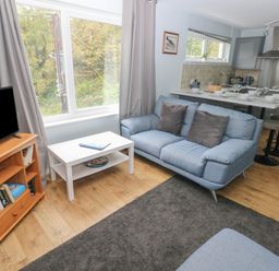 A living room with a blue sofa a white coffee table a wooden tv stand with a television and books a kitchen area with a breakfast bar and stools at Kittiwake Apartment 22 in Little Haven near Broad Haven