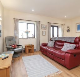 A living room with a red sofa gray armchair wooden tables and wall clock at Badgers Sett in Stibb near Kilkhampton
