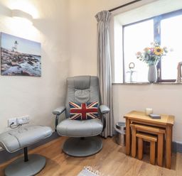A corner with a gray chair and matching footrest with a union jack pillow a set of nested wooden tables and a window with flowers and lanterns at Badgers Sett in Stibb near Kilkhampton