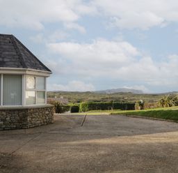 A driveway next to a house with stone wall and large windows overlooking a grassy landscape with hills and a street lamp at Horn Head View in Carrigart County Donegal