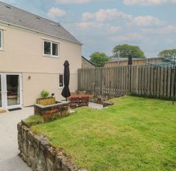 A backyard with a lawn enclosed by a wooden fence and patio furniture with umbrellas at The Packing House in Rosudgeon