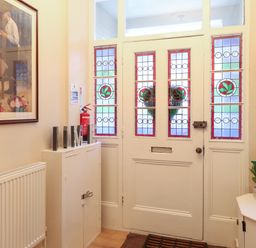 An entryway with stained glass front door a small cabinet a radiator a framed painting a plant on a corner shelf and a fire extinguisher at Grosvenor House in Kirkley Rise