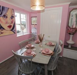 A dining room with a wooden table set for four with pink dishes and a large portrait on the pink wall at Cherry Blossom Cottage in Stratford-Upon-Avon